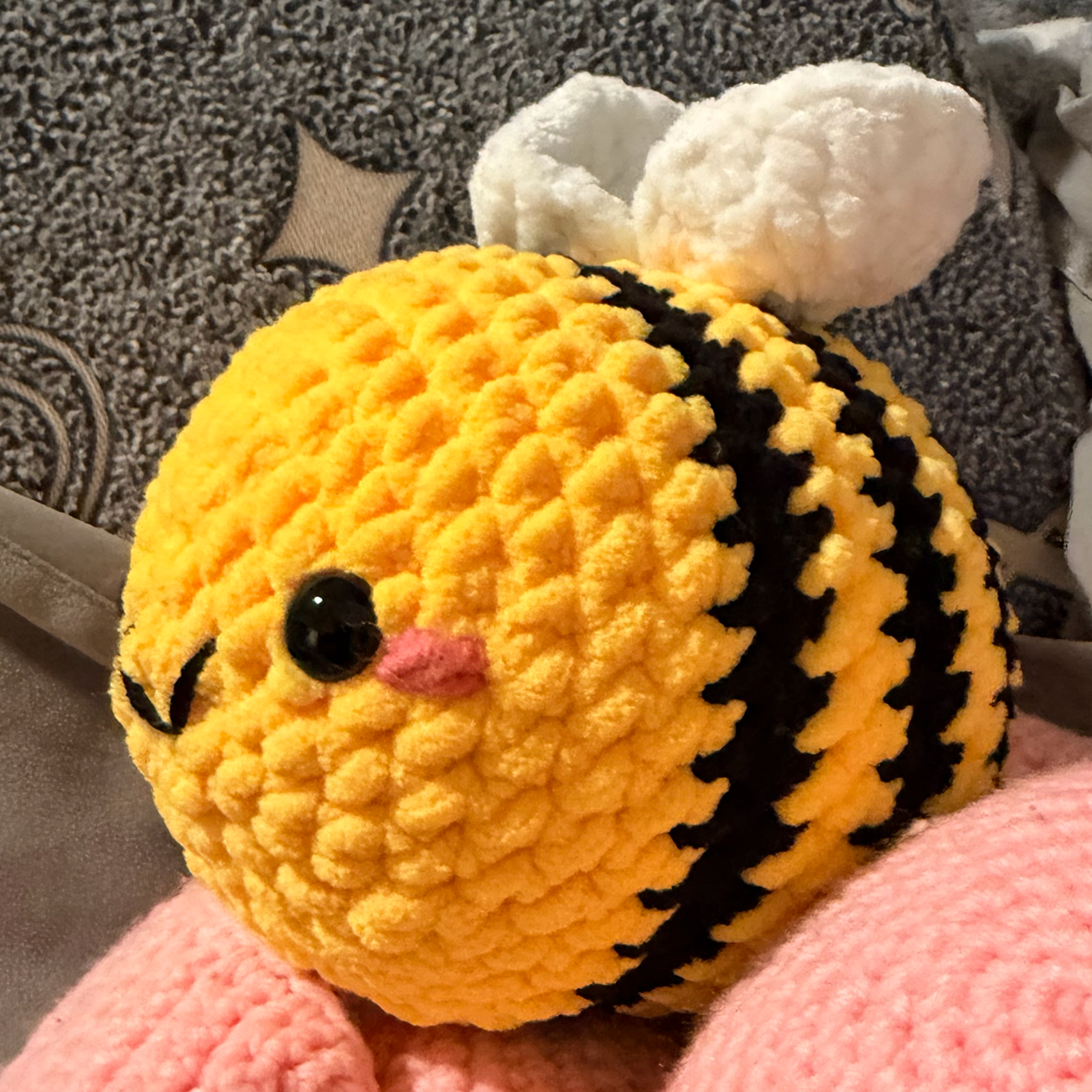 Crocheted bee plush toy on a pink blanket with a patterned pillow in the background